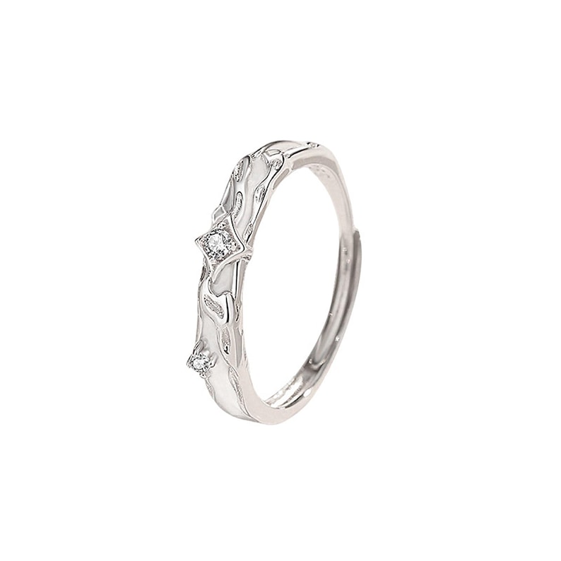 K&ouml;nnte beinhalten: Silberfarbener Ring mit einem dekorativen wei&szlig;en Design und zwei kleinen, klaren Edelsteinen. Der Ring hat ein offenes Design und ist wahrscheinlich verstellbar. Das dekorative Design hat ein flie&szlig;endes, organisches Muster.