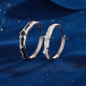 K&ouml;nnte beinhalten: Zwei silberne Ringe mit schwarz-wei&szlig;en Emaille-Details. Ein Ring hat ein schwarzes rautenf&ouml;rmiges Design, der andere ein wei&szlig;es Design mit einem kleinen, klaren Edelstein. Die Ringe sind vor einem dunkelblauen Hintergrund platziert.