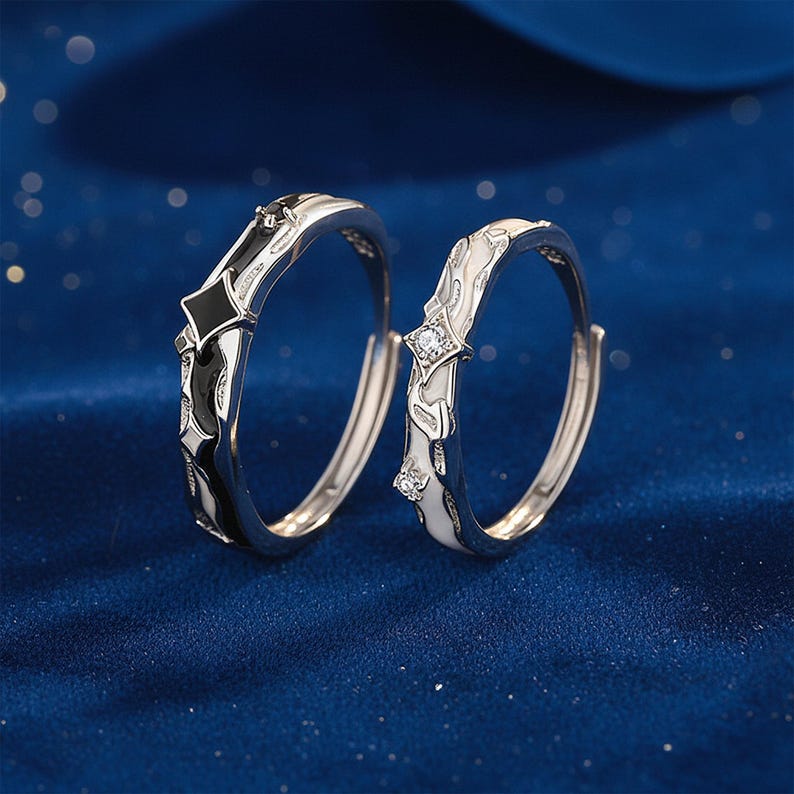 K&ouml;nnte beinhalten: Zwei silberne Ringe mit schwarz-wei&szlig;en Emaille-Details. Ein Ring hat ein schwarzes rautenf&ouml;rmiges Design, der andere ein wei&szlig;es Design mit einem kleinen, klaren Edelstein. Die Ringe sind vor einem dunkelblauen Hintergrund platziert.