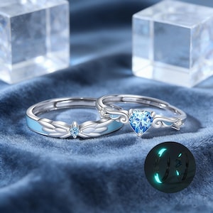 May include: Two silver rings with blue accents and gemstones. One ring features a heart-shaped blue gemstone, while the other has a smaller round blue gemstone. The rings have a light blue enamel design and are set against a blue velvet background.