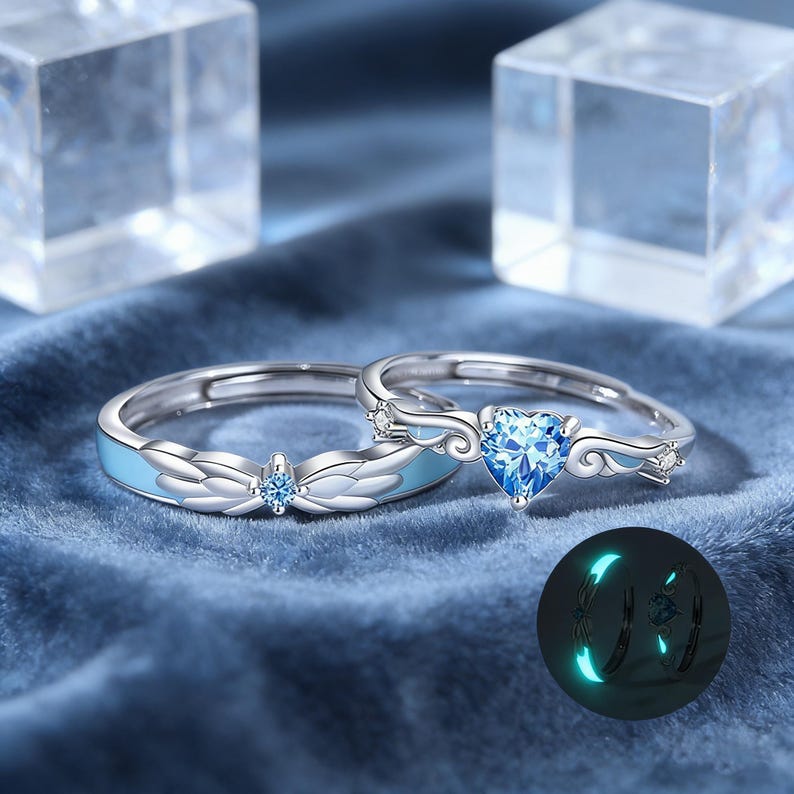 May include: Two silver rings with blue accents and gemstones. One ring features a heart-shaped blue gemstone, while the other has a smaller round blue gemstone. The rings have a light blue enamel design and are set against a blue velvet background.