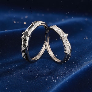 K&ouml;nnte beinhalten: Zwei silberne Ringe mit schwarz-wei&szlig;en Emaille-Details. Ein Ring hat einen diamantartigen Akzent. Die Ringe sind vor einem dunkelblauen Hintergrund mit funkelnden Highlights platziert, was auf einen romantischen oder besonderen Anlass hindeutet.