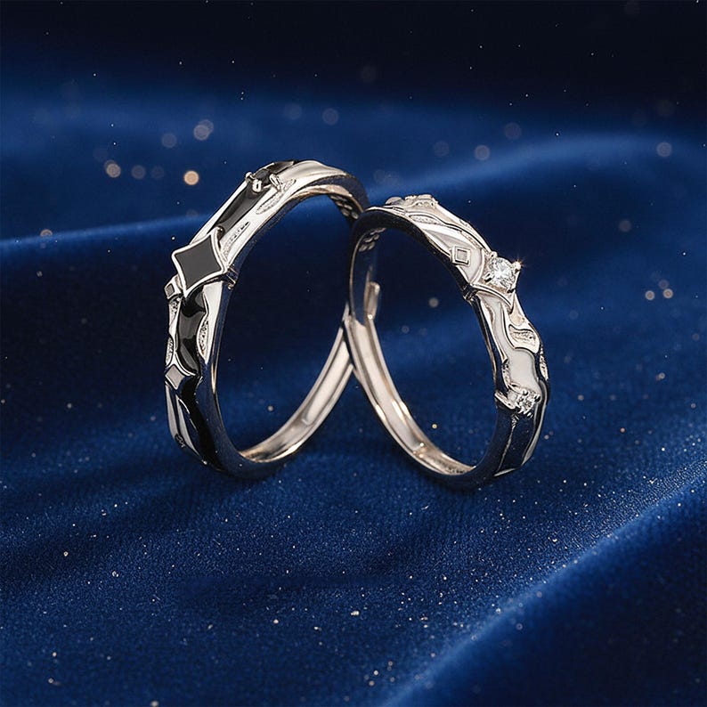 K&ouml;nnte beinhalten: Zwei silberne Ringe mit schwarz-wei&szlig;en Emaille-Details. Ein Ring hat einen diamantartigen Akzent. Die Ringe sind vor einem dunkelblauen Hintergrund mit funkelnden Highlights platziert, was auf einen romantischen oder besonderen Anlass hindeutet.