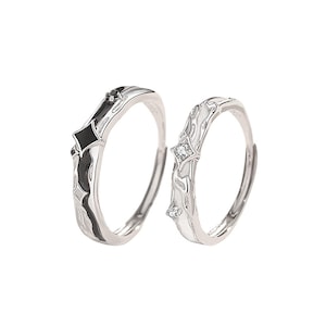 K&ouml;nnte beinhalten: Zwei silberne Ringe mit einzigartigen Designs. Ein Ring hat ein schwarz-silbernes geometrisches Muster, der andere ein wei&szlig;-silbernes Blumenmuster mit kleinen, funkelnden Steinen. Die Ringe sind vor einem schlichten wei&szlig;en Hintergrund platziert.