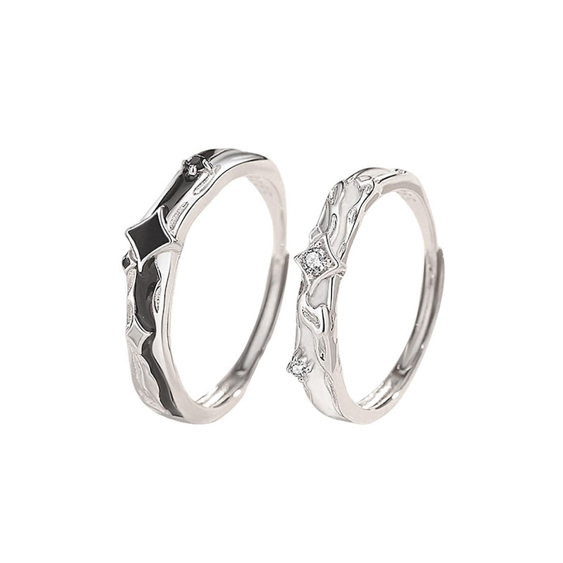 K&ouml;nnte beinhalten: Zwei silberne Ringe mit einzigartigen Designs. Ein Ring hat ein schwarz-silbernes geometrisches Muster, der andere ein wei&szlig;-silbernes Blumenmuster mit kleinen, funkelnden Steinen. Die Ringe sind vor einem schlichten wei&szlig;en Hintergrund platziert.