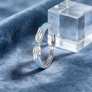 May include: A silver ring with a light blue enamel band and a small, round, blue gemstone. The ring features a stylized wing design on either side of the gemstone. The ring is set against a blue velvet background.