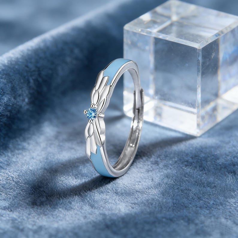 May include: A silver ring with a light blue enamel band and a small, round, blue gemstone. The ring features a stylized wing design on either side of the gemstone. The ring is set against a blue velvet background.