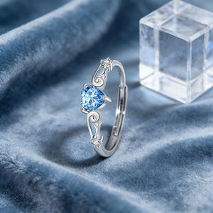 May include: A silver ring with a heart-shaped, light blue gemstone. The ring features decorative flourishes and small clear stones. The ring is set against a blue velvet background.