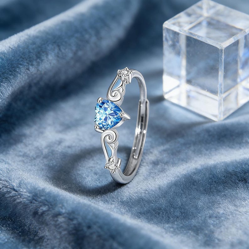May include: A silver ring with a heart-shaped, light blue gemstone. The ring features decorative flourishes and small clear stones. The ring is set against a blue velvet background.