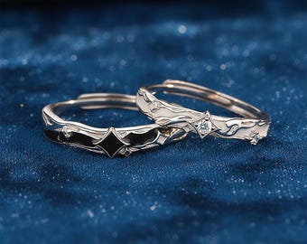 May include: Two silver adjustable rings. One ring features a black enamel design with a diamond-shaped accent. The other ring has a white enamel design with a small clear gemstone. Both rings are on a blue background.