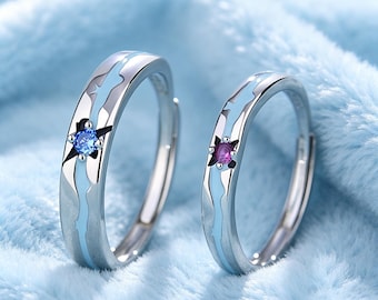 May include: Two silver rings with a light blue and white design. Each ring features a star-shaped accent with a central gemstone, one blue and one purple. The rings are set against a soft, light blue background.