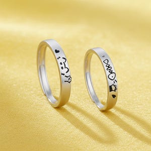 May include: Two silver rings with black etched designs. One ring features a heart, a cat face, and a paw print. The other ring has a fish bone, a cat face, a heart, and a paw print. The rings are displayed on a yellow surface.