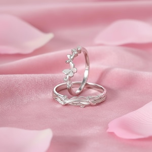 Puede incluir: Dos anillos de plata con diseños de mariposas y hojas. Un anillo presenta una mariposa y pequeñas piedras transparentes. El otro anillo tiene un diseño de hojas y vides. Los anillos están sobre un fondo rosa.