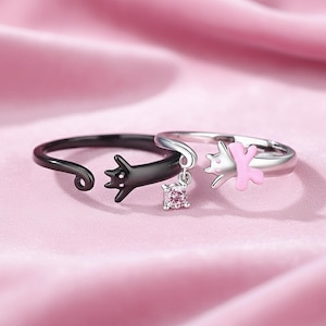 May include: Two adjustable cat rings. One is black with a dangling square pink gem, the other is silver with a pink cat design. The rings are on a pink satin surface. Jewellery.