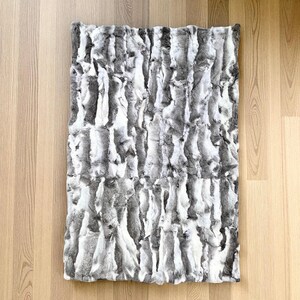 Può includere: Una coperta rettangolare in finta pelliccia nei toni del grigio e del bianco, con un motivo a righe verticali. La coperta è appoggiata su una superficie di legno chiaro.