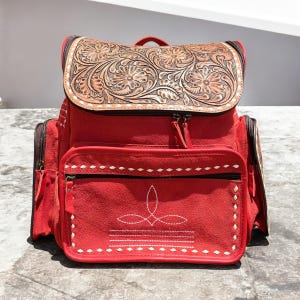 May include: Red suede backpack with a brown leather flap featuring floral tooling. The backpack has two front zippered pockets, side pockets, and white stitching details. The top handle and straps are also red.