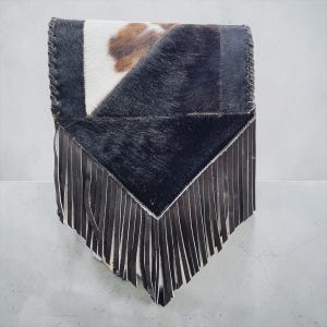May include: A cowhide clutch with a patchwork design. The top section features a mix of black, white, and brown fur, while the bottom has a black leather fringe. The edges are stitched with a thick thread.