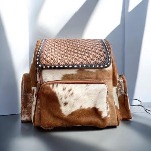 May include: A brown and white cowhide backpack with a decorative, embossed leather top flap. The backpack has a front pocket and two side pockets. The bag is made of leather and has a western style.