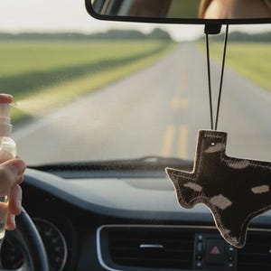 May include: A clear plastic spray bottle emitting a fine mist and a hanging car air freshener shaped like the state of Texas, featuring a dark brown and white cowhide pattern with visible stitching along the edges, suspended by a black cord.