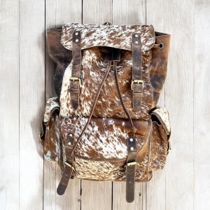 May include: A brown and white cowhide backpack with leather straps and brass buckles. The backpack has a flap closure and two side pockets. The bag is made of a natural material and has a rustic, outdoorsy aesthetic.