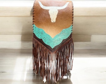 May include: A brown leather bag featuring a white cow skull graphic on the upper portion. The lower section is accented with a turquoise floral embossed leather overlay. The bottom edge is finished with long, thin brown leather fringe tassels.