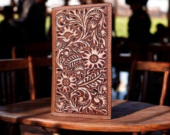 May include: A brown leather wallet with a floral tooled design. The wallet is sitting on a wooden table.