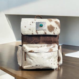 May include: A cowhide backpack with a brown leather body and a flap closure. The flap features a silver-colored decorative plate with a turquoise stone. The backpack has a front pocket and a top handle.