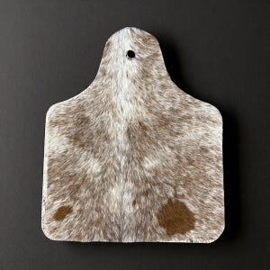 May include: A rectangular cutting board with a handle, featuring a natural cowhide pattern in shades of brown and white. The board has a hole near the handle for hanging. The cowhide pattern is visible across the entire surface.