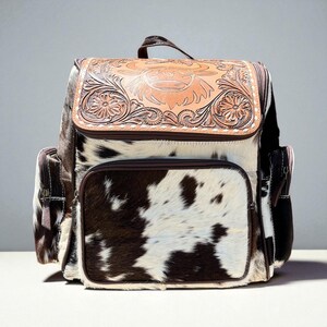 May include: A cowhide backpack with a brown leather flap featuring floral and bull carvings. The backpack has a brown and white spotted pattern, with brown leather trim and a top handle. It has two side pockets and a front pocket.