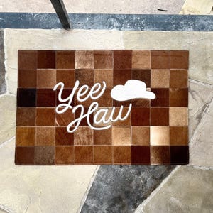 May include: Brown and tan cowhide doormat with a checkerboard pattern. The white text "Yee Haw" and a cowboy hat are centered on the mat. The mat is rectangular.