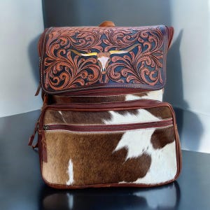 May include: A western-style backpack with a brown and white cowhide lower section and a tooled leather upper section. The upper section features a detailed floral design and a longhorn skull. The backpack has multiple zippered compartments.
