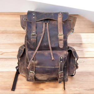 May include: A brown leather backpack with multiple pockets and adjustable straps. The backpack features a flap closure with buckles and a drawstring top. The leather has a distressed finish, giving it a vintage look. The bag is designed for carrying items.