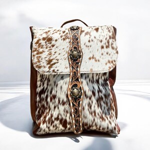 May include: A cowhide backpack with brown and white spotted fur. The bag features a brown leather base and a decorative strap with metal accents. The flap closure and handle are also brown leather.