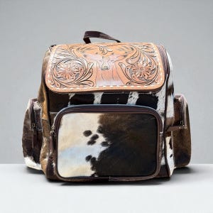 May include: A western-style backpack with a brown leather flap featuring a carved floral and deer design. The body of the backpack is made of cowhide with brown, black, and white patches. It has a top handle, side pockets, and a front zippered pocket.