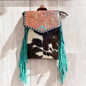 May include: A western-style handbag with a brown leather flap featuring floral tooling and turquoise accents. The bag's body is made of cowhide with black and white patches, and it has turquoise fringe.