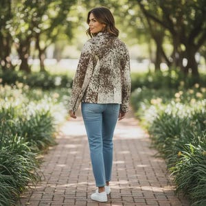 May include: A woman wearing a brown and cream-colored patterned jacket and light blue jeans walks on a brick pathway. The jacket has a zip-up front and long sleeves. White sneakers complete the outfit.