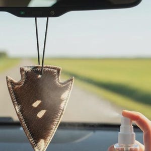 May include: An arrowhead-shaped car air freshener with a brown and white cowhide pattern, suspended by a black cord from a rearview mirror, being sprayed with a clear liquid from a small, handheld, transparent plastic spray bottle.