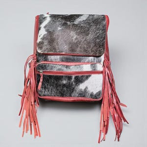 May include: A red leather backpack with a cowhide flap and front pocket. The backpack features long red fringe on the sides and a zipper closure. The cowhide has a black and white pattern.