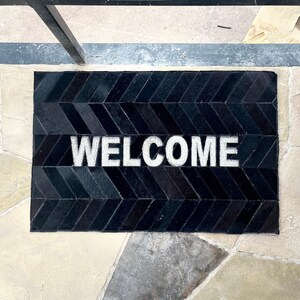 May include: A rectangular black doormat with a chevron pattern and the white word "WELCOME" in the center. The mat is made of a textured material and sits on a stone tile floor.