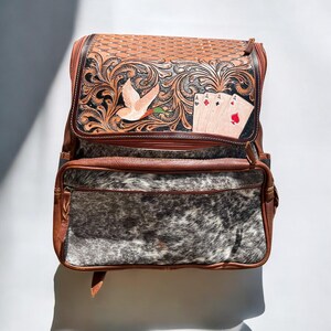May include: Brown leather backpack with a decorative floral and bird design on the flap. The backpack features a front pocket with a cowhide pattern and a top flap with a woven texture. The flap also has playing cards.