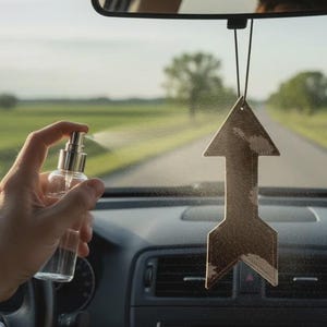 Puede incluir: Un frasco pulverizador de vidrio transparente con boquilla plateada rocía un ambientador colgante de color marrón con forma de flecha. El ambientador está suspendido por un hilo negro fino del espejo retrovisor dentro de un vehículo.