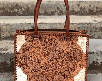 May include: A brown leather tote bag with a white and brown cowhide pattern. The bag has a floral design embossed into the leather. The bag has two top handles.