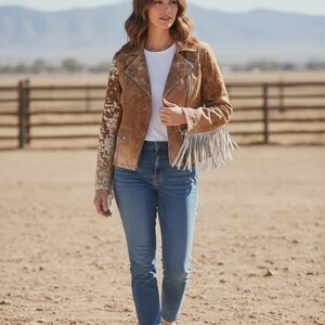 May include: A woman wearing a brown suede jacket with cowhide print sleeves and fringe detailing. She is wearing a white t-shirt, blue jeans, and white sneakers. The background features a wooden fence and mountains.