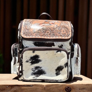May include: A cowhide leather backpack with a brown leather top flap featuring a carved design of a horse and floral elements. The backpack has a black and white spotted pattern, with two side pockets and a front pocket.