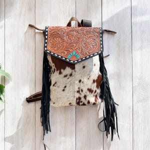 May include: A western-style backpack with a brown and white cowhide body and a tooled leather flap. The flap features floral designs, turquoise accents, and black fringe. The bag has brown leather straps and a handle.