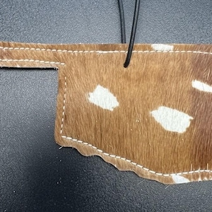 Puede incluir: Un adorno de piel de vaca marrón y blanco con la forma del estado de Oklahoma. La pieza presenta una superficie de pelo texturizado con manchas blancas irregulares, rematada con costuras perimetrales blancas y un cordón negro para colgar.