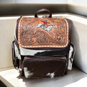 May include: A backpack featuring a tan leather flap with intricate floral tooling and a central graphic of a person riding a spotted horse. The body is constructed from black and white cowhide with brown leather trim, zippers, and a top handle.