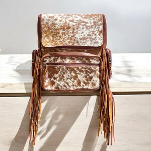 May include: A brown and white cowhide backpack featuring a large top flap and a front zippered pocket. The bag is accented with long, thin leather fringe tassels hanging from both sides of the front compartment and includes side pockets.