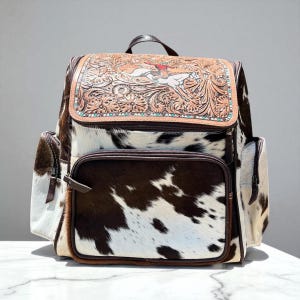 May include: A cowhide backpack with a brown leather flap featuring a carved design of a rider on a horse. The backpack has a brown and white spotted pattern, with brown leather trim and pockets. The bag has a top handle and two side pockets.