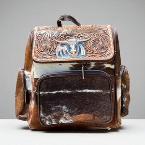 May include: A brown and white cowhide backpack with a decorative embossed leather flap featuring a bull design. The backpack has a front zippered pocket and side pockets. The bag has a top handle and adjustable straps.